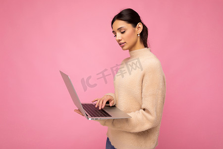侧面照片：迷人漂亮的年轻女士拿着笔记本电脑，隔离在墙壁背景上，看着上网本，在键盘上打字，思考着什么