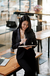 在办公室工作并记笔记的亚洲女商人肖像，坐在办公桌上使用笔记本电脑、平板电脑的女性