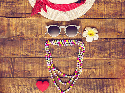 平躺的帽子、项链、太阳镜、花朵和红心形作为旅行者的配饰，在带有复制空间的木制背景下度过暑假。