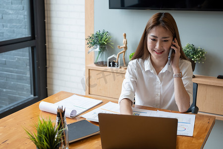 月度工作报告摄影照片_微笑的亚洲女性在笔记本电脑上工作时接听手机电话，获取财务会计号码、文书统计月度报告