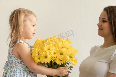 母亲、母亲节、生日、童年、家庭观念 — 身穿蓝色彩色连衣裙的特写小女孩祝贺并给年轻母亲明亮的黄色雏菊花束。白色背景。