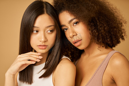 两位美丽的混血年轻女性的脸部特写，她们皮肤完美，站在米色背景中，在镜头前摆出姿势