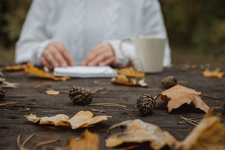 身穿白色毛衣的年轻女子坐在公园一张黑色的旧桌子旁，端着一杯加咖啡的茶，在黄色秋叶的背景下读书写字。