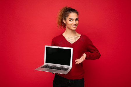 美丽迷人的迷人年轻卷发黑金发女士穿着红色毛衣站在红墙背景上孤立的侧面肖像照片，拿着电脑笔记本电脑，上面有空的复制空间模型，指向上网本显示器和键盘厕所