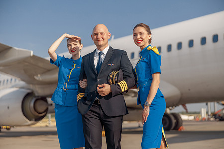 快乐的飞行员和两位漂亮的空姐一起站在飞机前，着陆后微笑