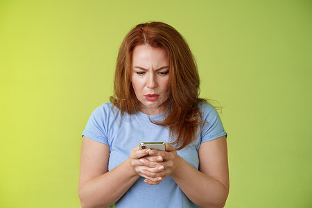 困惑而不确定的红发中年女性学习如何使用社交媒体试图理解表情符号看起来强烈聚焦的智能手机显示屏阅读重要新闻消息站绿色背景