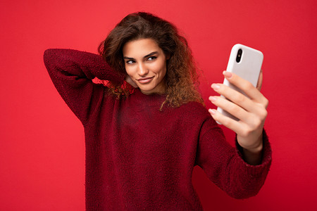 美丽的年轻女子，卷发，穿着深红色毛衣，隔离在红色背景墙上，拿着和使用智能手机，看着电话屏幕，自拍