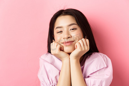 梦幻浪漫的亚洲女孩的特写肖像，手上瘦着脸，微笑着，面带钦佩和幸福，站在粉红色的背景下