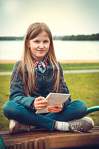 长木凳摄影照片_一个金发女孩坐在公园的木凳上，面对蓝色的大海，写了一封信
