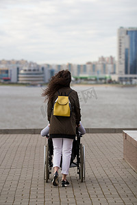 关爱残疾人摄影照片_码头上年轻女子与轮椅上的残疾人同行的后视图