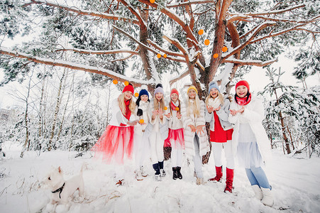 一大群拿着橘子的女孩站在冬天的森林里。白雪皑皑的森林里，穿着红色和白色衣服、带着水果的女孩