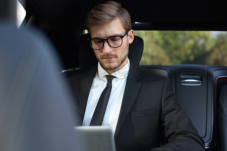 全套西装摄影照片_英俊自信的商人穿着西装，坐在车里用智能手机交谈，并使用笔记本电脑工作。
