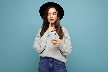 美丽的黑发年轻女子戴着黑帽子和灰色毛衣，手持智能手机，低头看着背景中孤立的电话短信，思考着