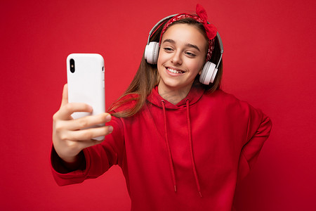 身穿红色连帽衫、身穿红色背景隔离的黑发美女微笑的特写镜头，手持并使用智能手机拍摄自拍照片，戴着白色无线耳机，听着酷炫的音乐，看着手机显示屏