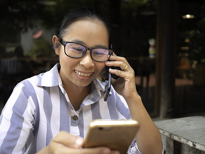 欢快的商务亚洲年轻女性手持智能手机上班，同时在咖啡厅打电话，脸上带着快乐和微笑。