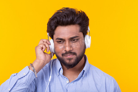 正在营业摄影照片_留着胡子的英俊轻松的印度男子正在黄色背景的耳机里听最喜欢的音乐。