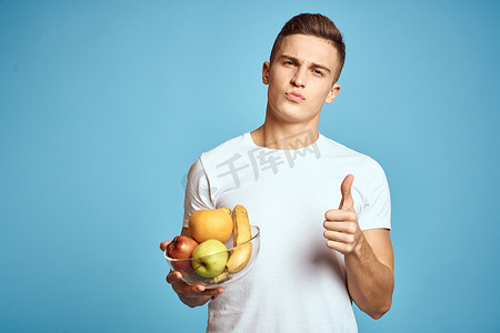 香蕉橙子摄影照片_快乐的男人，手里拿着新鲜水果，双手蓝色背景白色T恤维生素香蕉橙子苹果