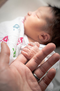 一个新生女婴在毯子襁褓中，用她的小手和手指包裹着父亲的手指，安然入睡。