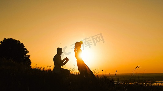情侣傍晚摄影照片_男人跪在高山上向女人求婚的剪影-情侣在日落时订婚-将戒指戴在女孩的手指上
