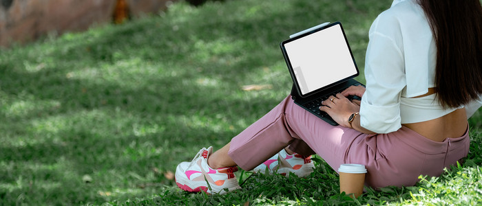特写学生与数字平板电脑空白屏幕坐在绿草草坪上，在笔记本上写作，教育技术，大学，高中，青少年概念。