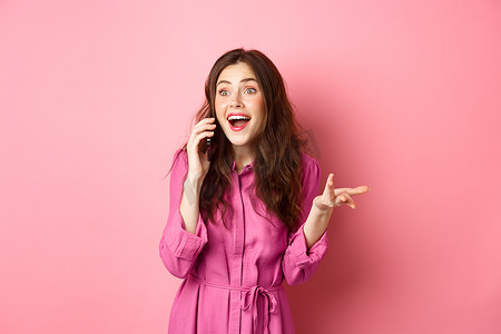 欢快的黑发女人在手机上说话，打手势和大笑，进行随意有趣的谈话，站在粉红色的背景上