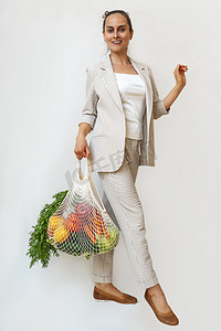 一位身穿浅色西装的女士拿着一个棉质购物袋和装有蔬菜的可重复使用的网状购物袋。