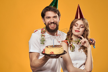 快乐的男人和女人，带着生日蛋糕生日帽派对黄色背景