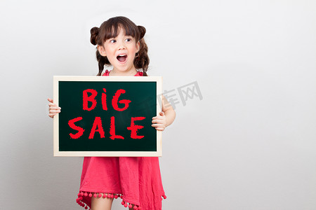 年终大促销红色摄影照片_年终概念购物中心促销中商品的大减价：可爱的女孩表演惊喜脸拿着粉笔标牌，上面写着红色的大减价。