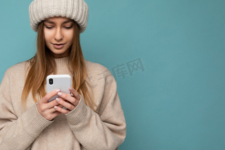 美丽的年轻金发女子的特写照片，穿着时尚的米色温暖毛衣和针织冬季温暖米色帽子，隔离在蓝色背景上，手握空白空间，使用手机短信查看智能手机显示 scr