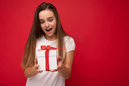 空礼盒摄影照片_照片中，美丽、积极、快乐的小女孩被隔离在红色的空地上，穿着休闲的白色 T 恤墙，拿着礼盒，玩得很开心