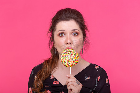 棒棒糖粉色背景摄影照片_糖果、对甜食的成瘾和情感概念 — 粉色背景中拿着棒棒糖的女人感到惊讶