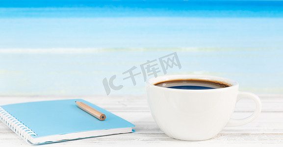 白色木桌上的咖啡杯和蓝色笔记本与明亮的大海