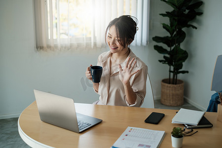 美丽的会计师坐在办公桌前的肖像，室内喝着热饮料，拿着咖啡杯，在视频会议时看着笔记本电脑。