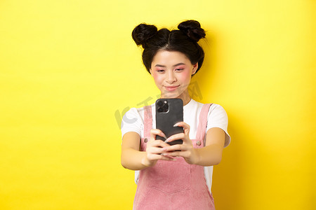 化妆小工具摄影照片_时尚的亚洲女孩在智能手机上拍照，用手机制作视频，微笑着，站在黄色背景上