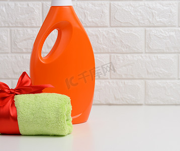 褶皱丝带摄影照片_浴室的白色架子上放着一条卷起的绿色毛巾，上面系着红色丝带，还有一瓶橙色塑料瓶，里面装着液体洗衣粉