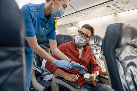 戴防护面罩的男子看着女空乘人员帮助他在飞机上调整并系紧安全带以确保安全旅行