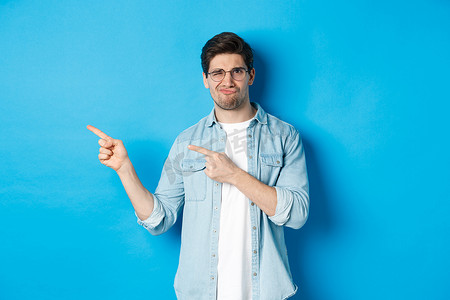 戴眼镜的不满和怀疑的男模特，用手指指着不好的东西，展示可怕的广告，站在蓝色背景上