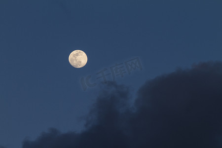 午夜的月亮摄影照片_满月在深蓝色的天空中升起，有些灰色的云