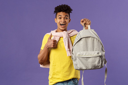 新学期摄影照片_快乐的年轻英俊男学生的肖像，男人推荐背包，拿着背包，竖起大拇指，为大学学期购买新设备，紫色背景
