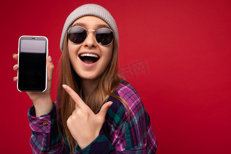 特写照片中，迷人的微笑积极的年轻金发女子穿着时尚的紫色衬衫和休闲的白色 T 恤灰色帽子和太阳镜，手持红色背景，展示手机，屏幕为空，用于观看凸轮的模型