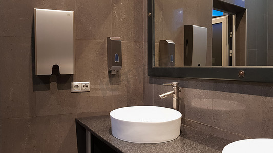 公共厕所里有白色圆形洗脸盆的镀铬水龙头，有大镜子和灰色墙壁，公共厕所的现代内部。