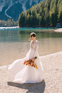 美丽的新娘穿着白色的袖子和蕾丝连衣裙，带着一束黄色的秋干花和牡丹玫瑰，在意大利的布拉伊斯湖上。
