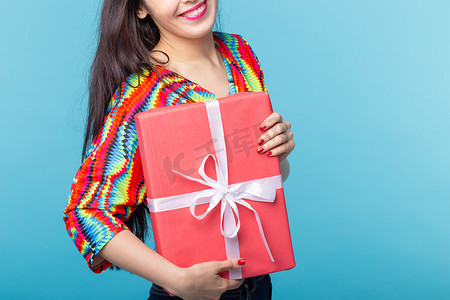 特写镜头：开朗漂亮的年轻黑发女人手里拿着一个红色礼品盒，但背景是蓝色的。