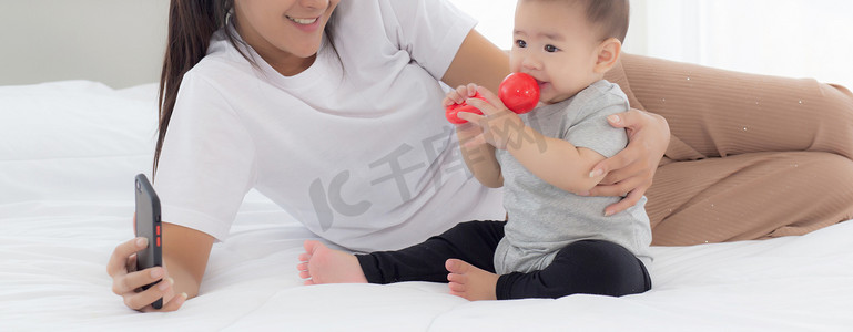 年轻的亚洲母亲和小女婴或新生儿在卧室的床上用智能手机自拍，幸福的母女在家里使用电话视频通话，两个人，家庭和沟通概念。