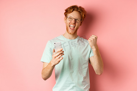 拿奖摄影照片_戴着眼镜和 T 恤的快乐红发男子赢得了网上奖，高兴和满意地大喊“是”，拿着智能手机，打拳头，粉红色背景