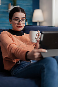 戴眼镜的学生坐在沙发上拿着杯子看着平板电脑显示屏。