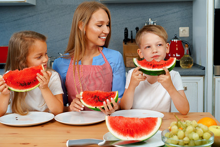 吃女孩西瓜摄影照片_甜蜜的家庭，母亲和她的孩子在厨房里吃西瓜玩得很开心