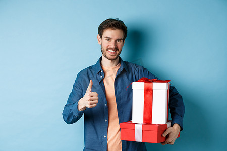 微笑的男人拿着浪漫的礼物，竖起大拇指，庆祝情人节，为爱人买礼物，站在蓝色背景上