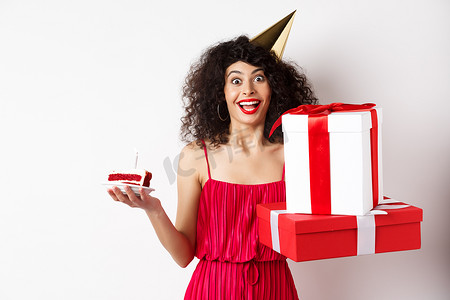 穿着派对帽和红色连衣裙的快乐女孩，庆祝生日，拿着惊喜礼物和生日蛋糕，享受假期，站在白色背景中