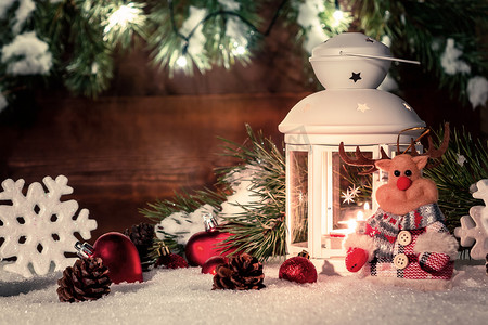 雪地上放着燃烧着蜡烛的白色灯笼，周围环绕着木墙、圣诞树枝和灯的圣诞装饰